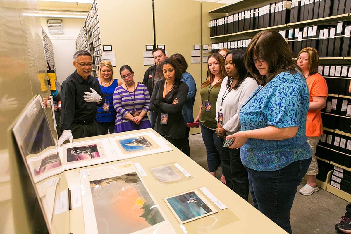 A group of people observing and discussing Disney Animation archival materials displayed on a table, guided by an individual wearing gloves.