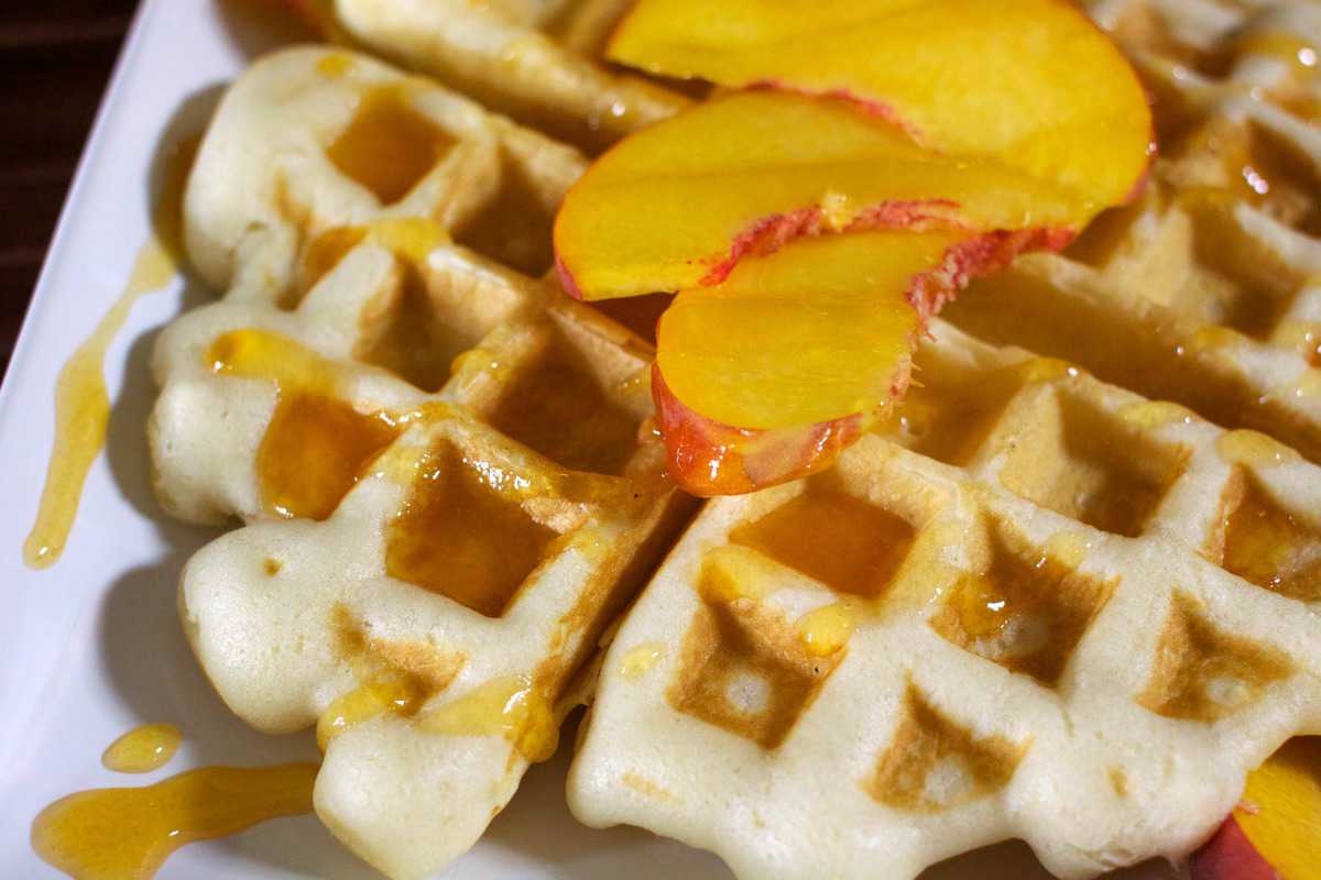 Belgian pancake topped with peach slices and drizzled with syrup on a white plate.
