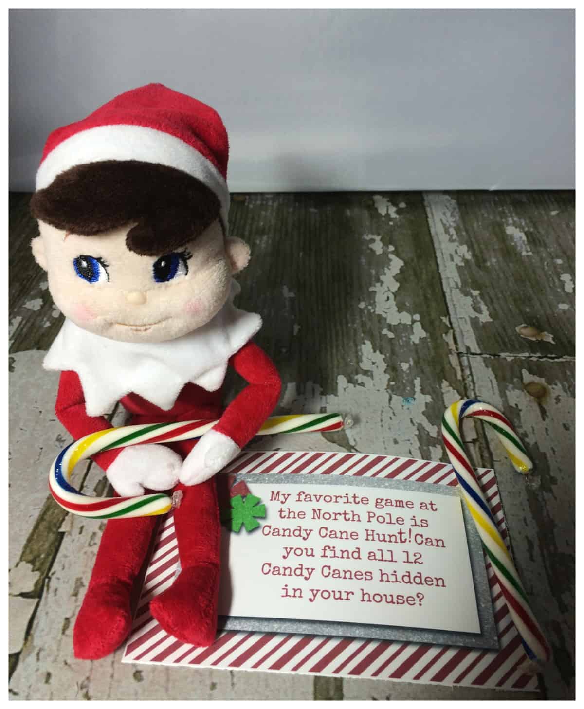 A plush elf toy with a Santa hat, resembling the "Elf on the Shelf" style, is seated next to a card and candy canes on a wooden surface, alongside printables available for free