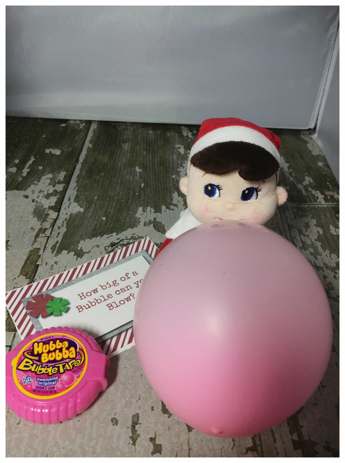 A plush elf on the shelf toy with a pink bubble gum balloon next to a Hubba Bubba container and a note that reads, "How big can a bubble get before it bursts?