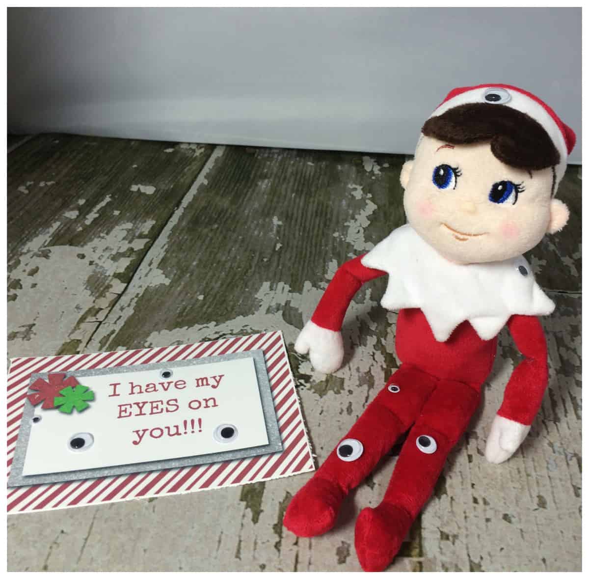 Plush elf doll sitting next to a playful note with the message "i have my eyes on you!!" against a weathered floor backdrop, accompanied by elf on the shelf printables free.