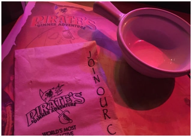 A souvenir program and a napkin from "Pirate's Dinner Adventure Buena Park" beside a white mug on a table illuminated by red ambient lighting.