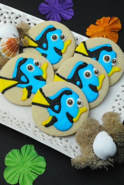 Plate of Finding Dory Sugar Cookies decorated with colorful icing to resemble tropical fish, surrounded by seashells and artificial sea-themed decorations.