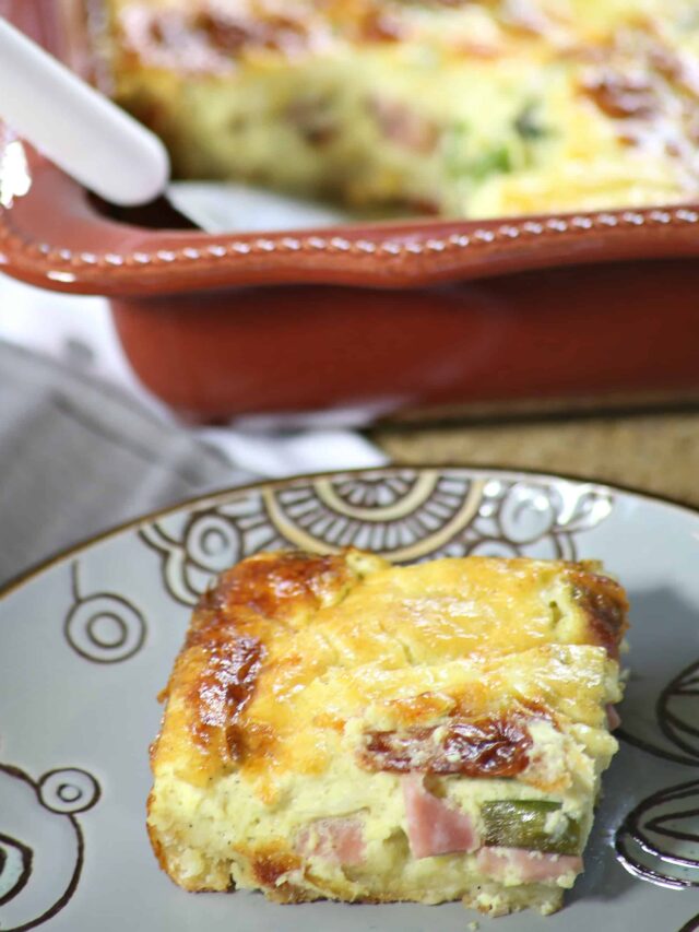 A close-up of a slice of Easy Ham and Cheese Quiche with green vegetables on a decorative plate. The casserole dish is visible in the background.