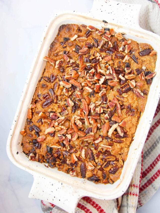 A baked dish filled with a nut-topped sweet potato casserole, possibly a bread pudding or casserole, inside a white rectangular baking dish. The dish is placed on a striped cloth.