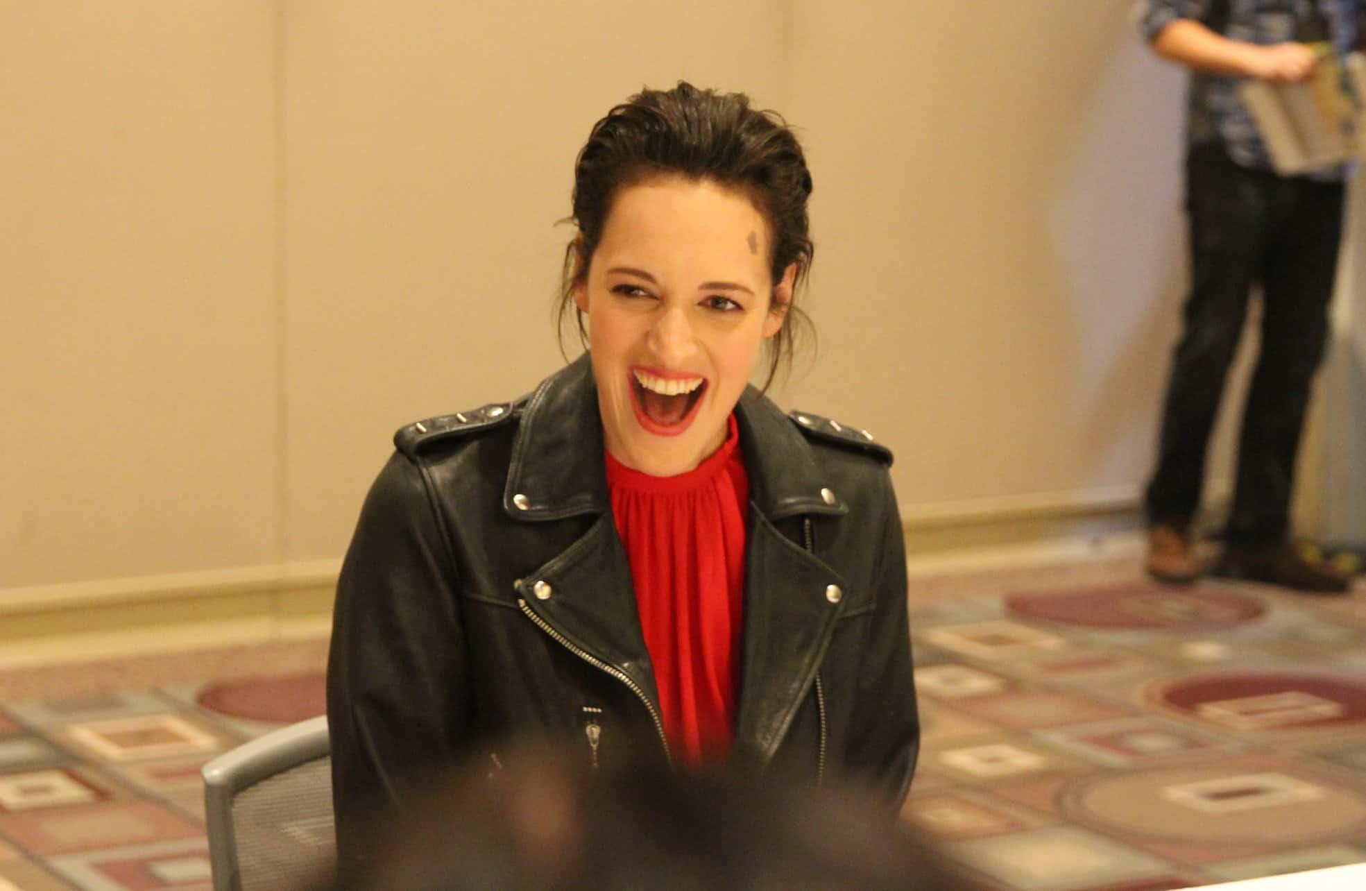Phoebe Waller-Bridge sitting at a table with a laptop and smiling at the camera