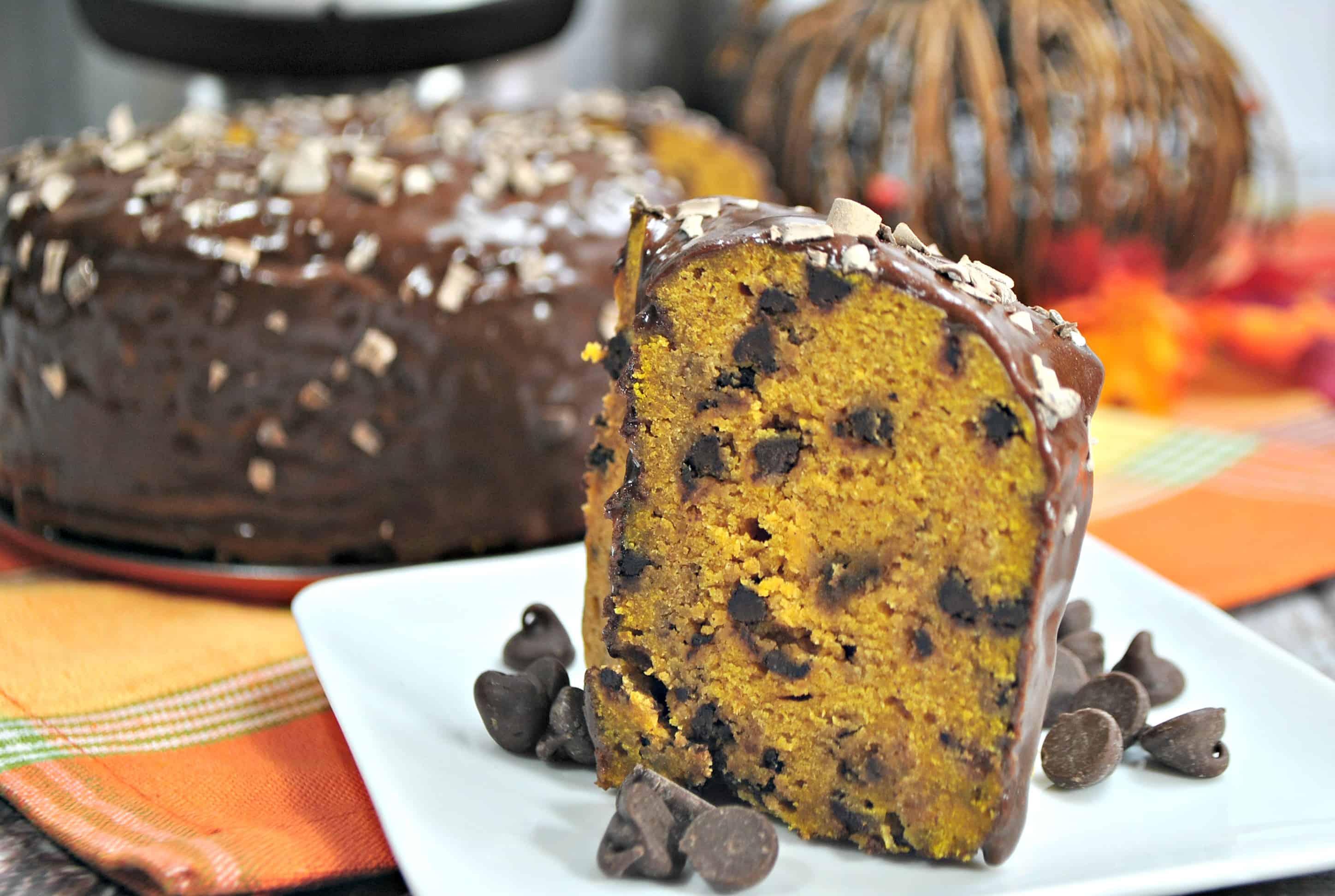 A slice of Instant Pot Pumpkin Chocolate Chip Cake with a glazed top, alongside more chocolate chips, with the remaining loaf in the background.