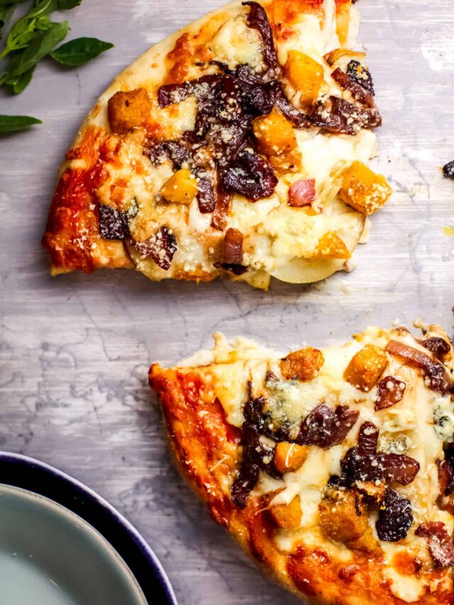 Two slices of pumpkin pizza on a light gray surface. One slice is slightly above the other, showcasing toppings like cheese, caramelized onions, and squash cubes. A plate is partially visible in the lower left corner.