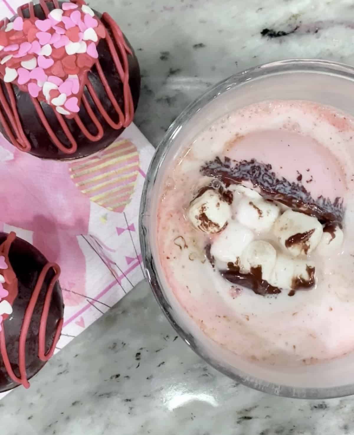 Two desserts on a marble surface: hot cocoa bombs with pink designs and a cup of strawberry milkshake topped with whipped cream and chocolate syrup.