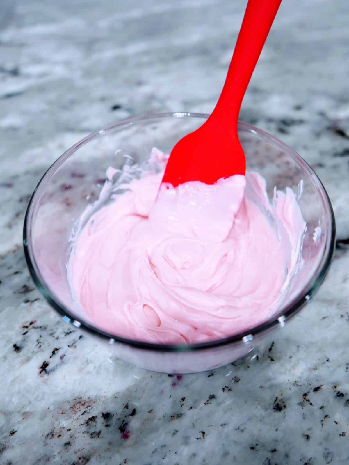 A red spatula mixing pink frosting in a clear glass bowl.
