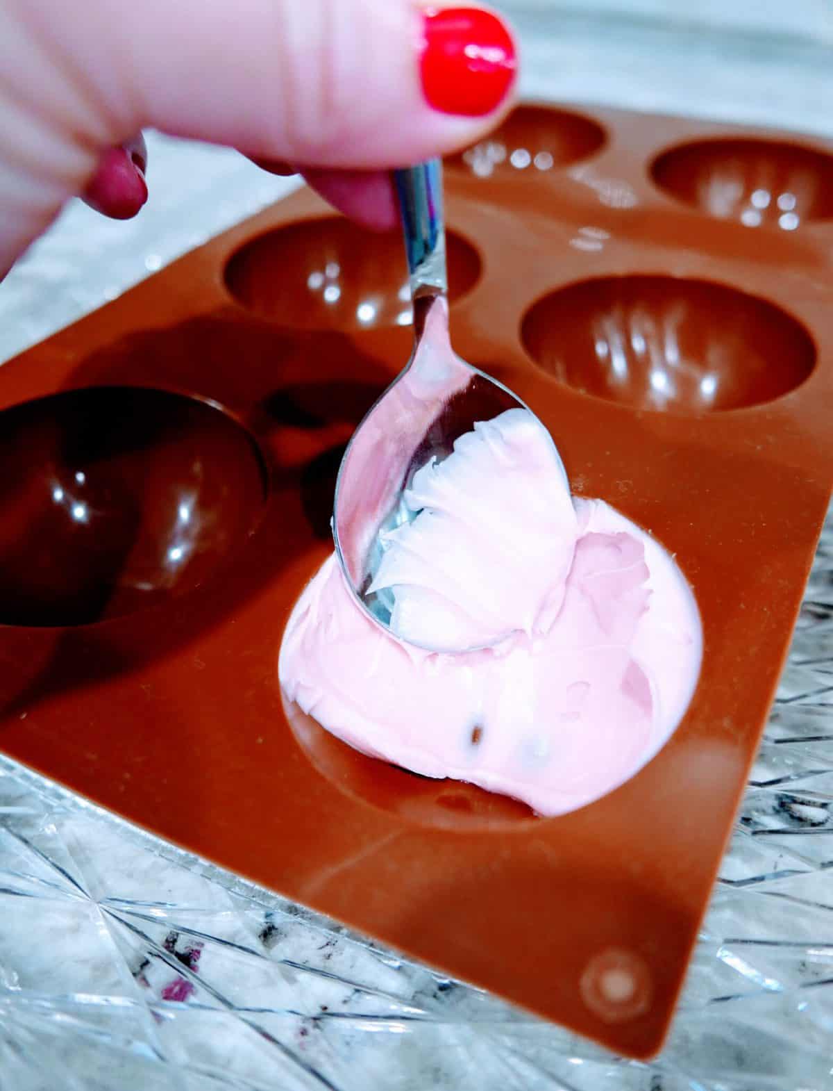 Filling a chocolate mold with pink mixture using a spoon.