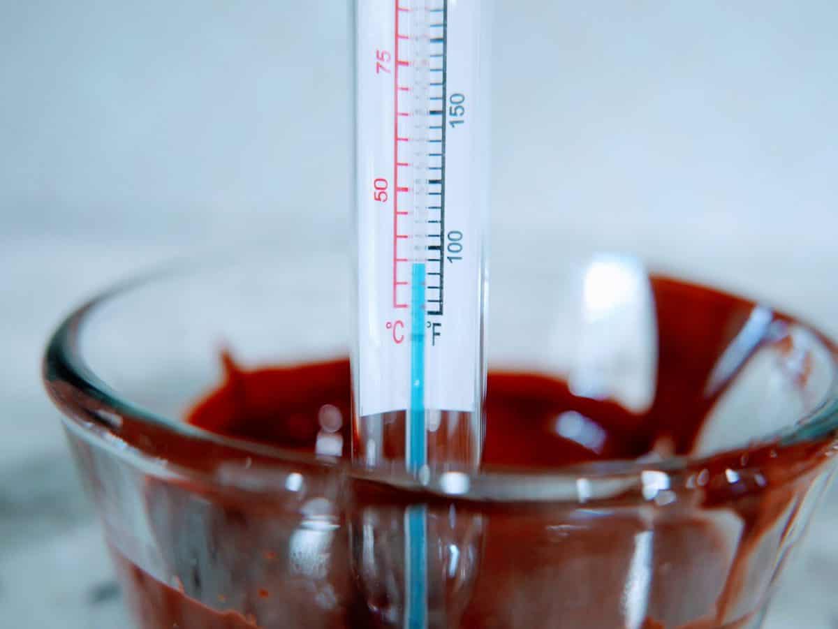 Candy thermometer measuring the temperature of melted chocolate in a glass bowl.