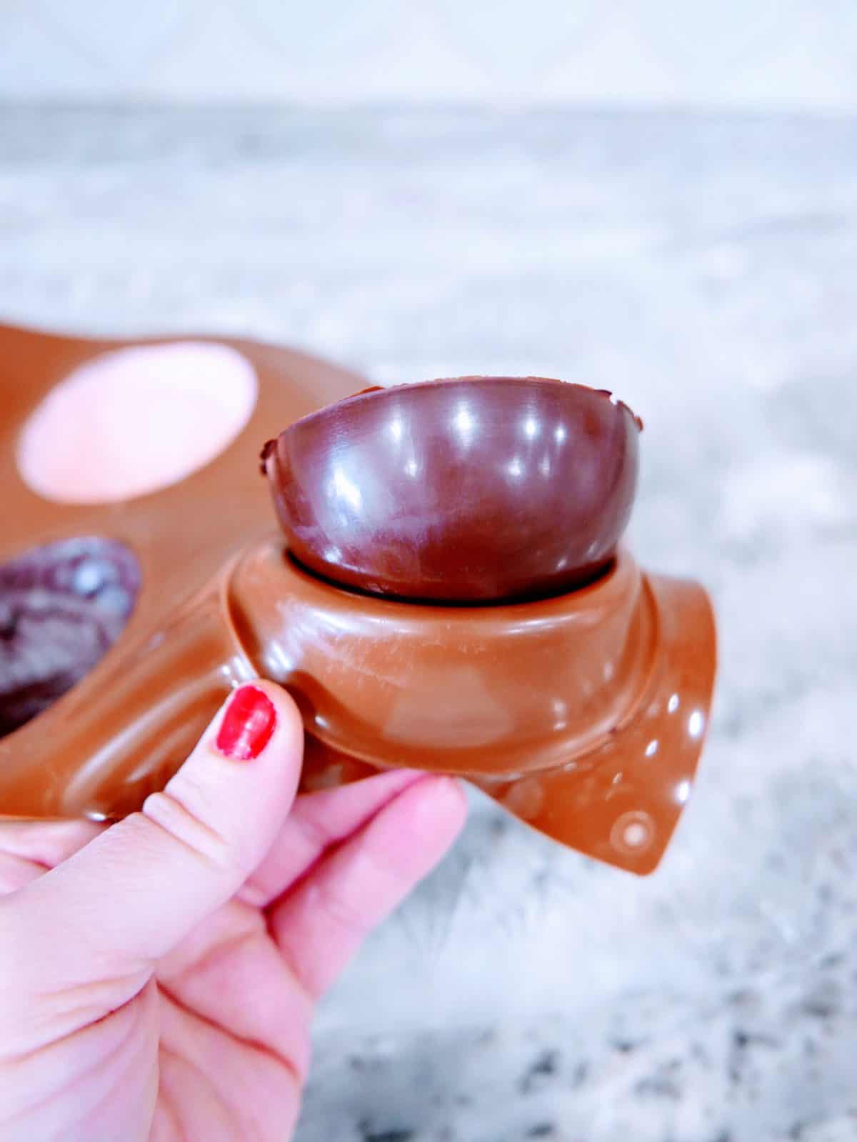A hand holding a half-open chocolate ball with a creamy filling, against a blurred background.