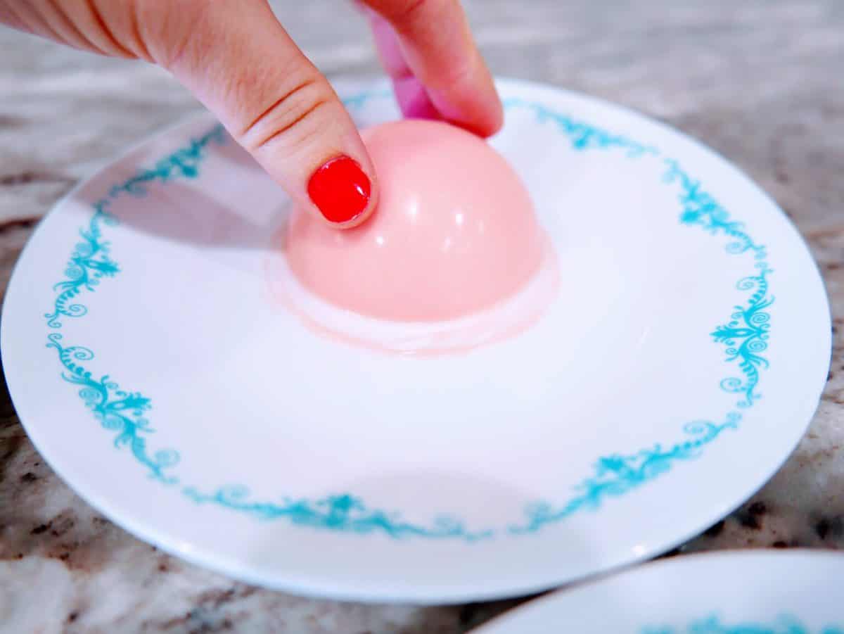 A person's hand with red nail polish pressing down on a pink dome-shaped gelatin dessert on a decorative plate.