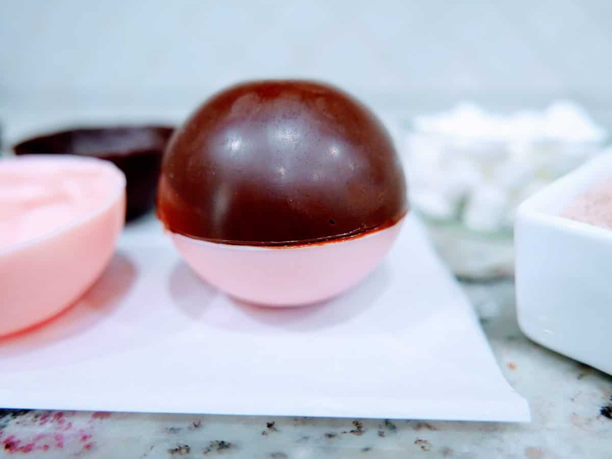 A chocolate sphere resting on a half-open pink plastic eggshell with blurred confectionery supplies in the background.