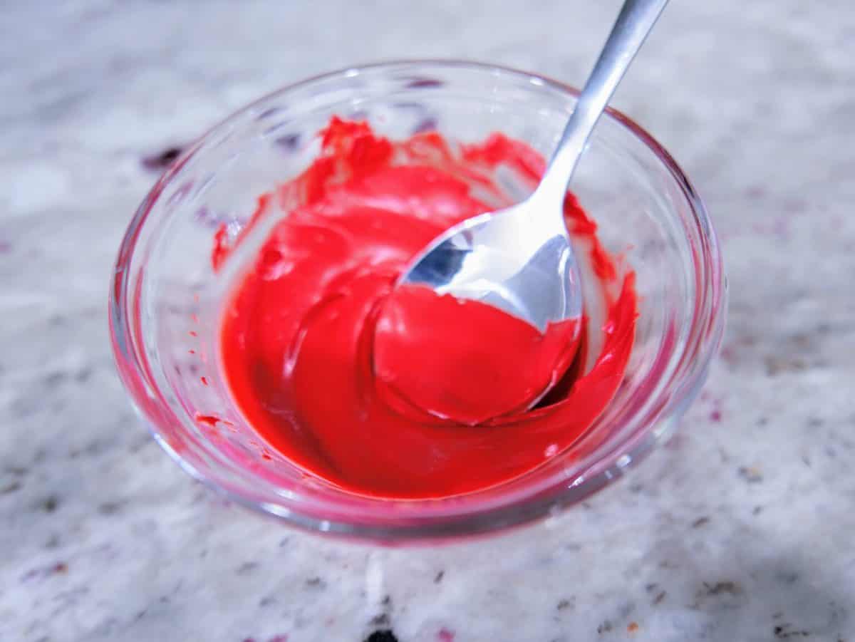 A bowl of red icing with a spoon on a marble surface.