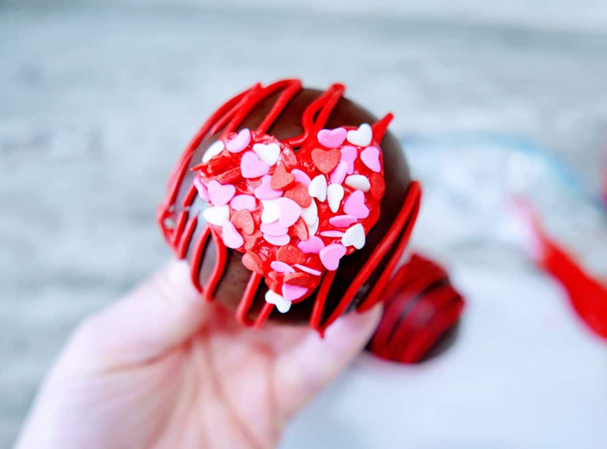 A hand holding a chocolate dipped strawberry adorned with red drizzle and colorful heart-shaped sprinkles.