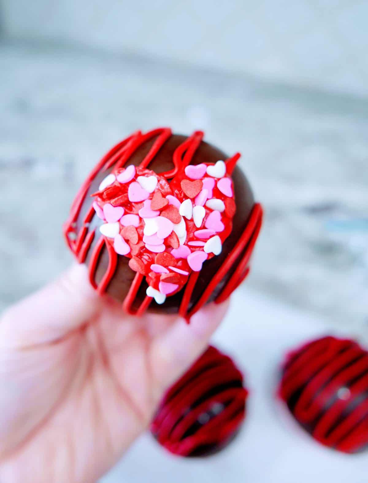 A hand holding a chocolate cupcake topped with red icing and heart-shaped sprinkles.