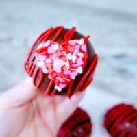 A hand holding a chocolate cupcake with red icing and heart-shaped sprinkles, inspired by a hot cocoa bombs recipe.