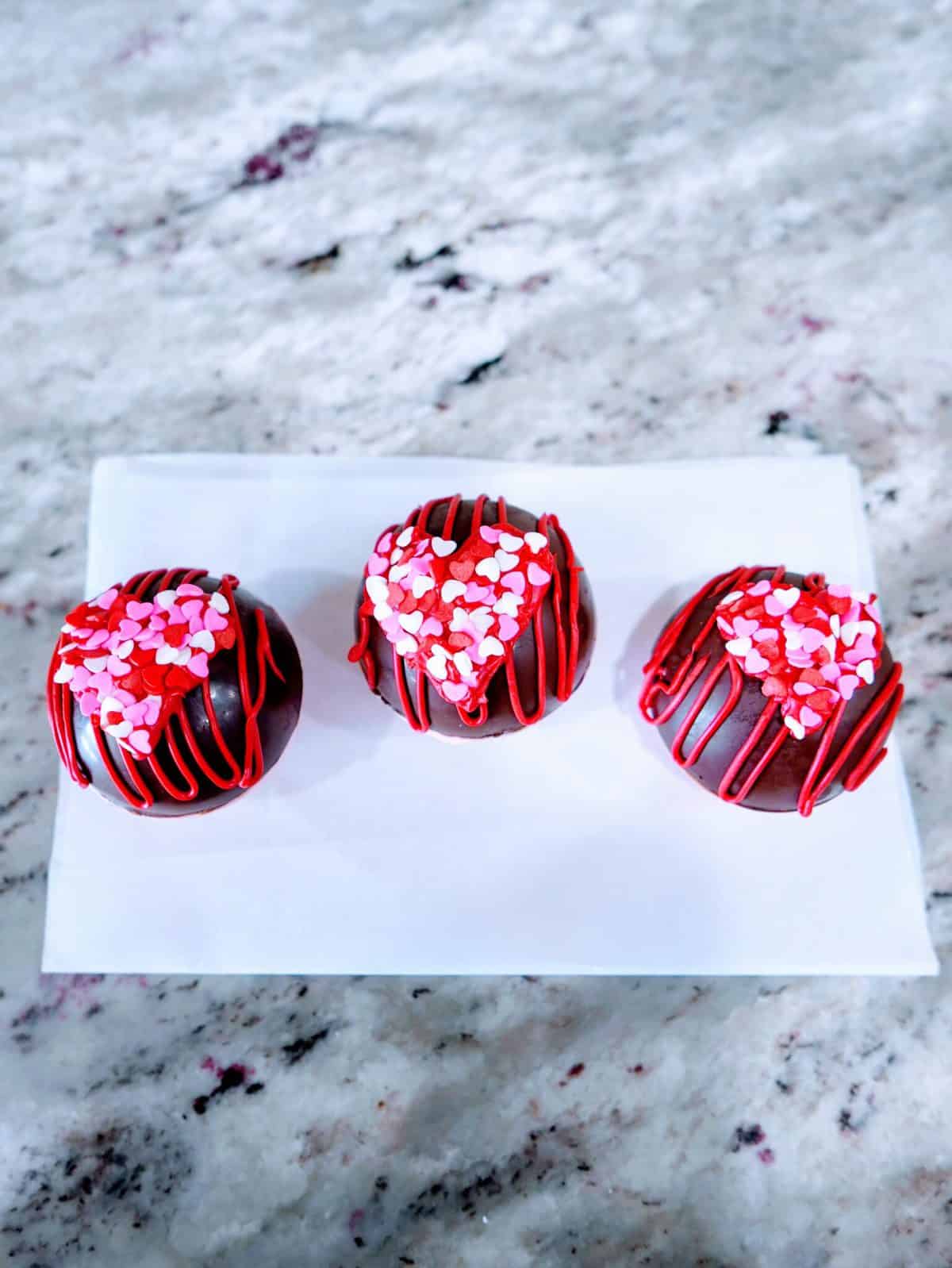 Three chocolate-covered strawberries with red drizzle and heart-shaped sprinkles on a white plate.