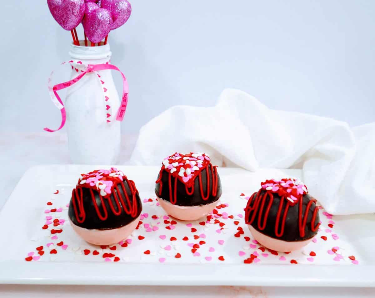 Three chocolate-covered strawberries with decorative icing and heart-shaped sprinkles, presented on a white plate with a festive bottle in the background.