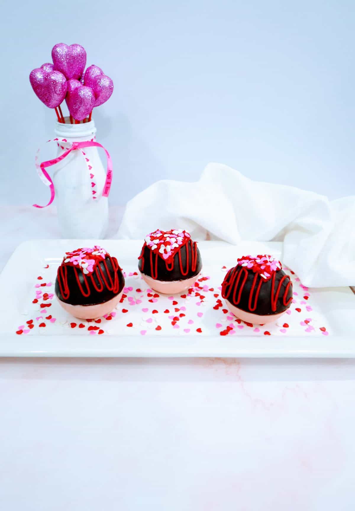 Three chocolate-covered desserts with red drizzle and sprinkles on a white plate, with a vase containing sparkly heart decorations in the background.