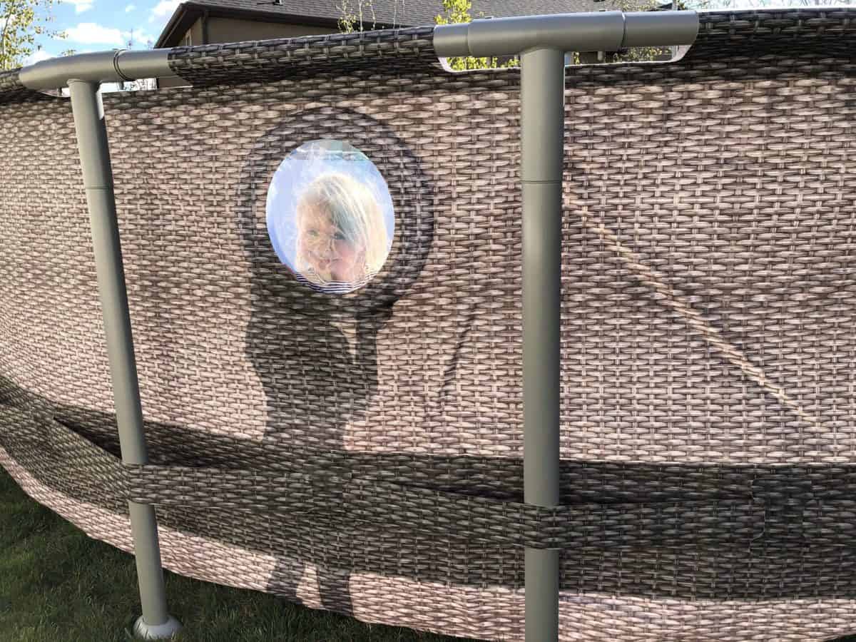 A child peering through a circular window in an outdoor wicker privacy screen.