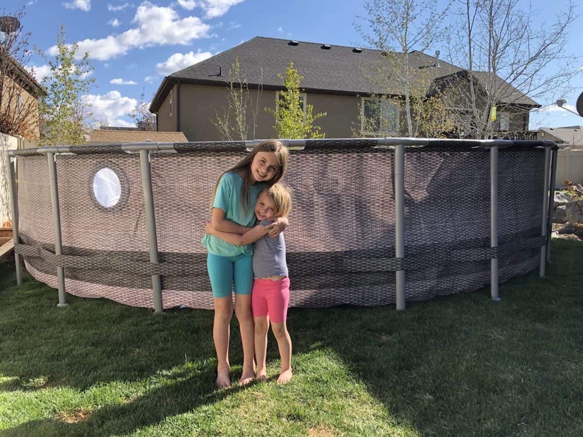 Two smiling children embracing in front of an above-ground pool in a sunny backyard.