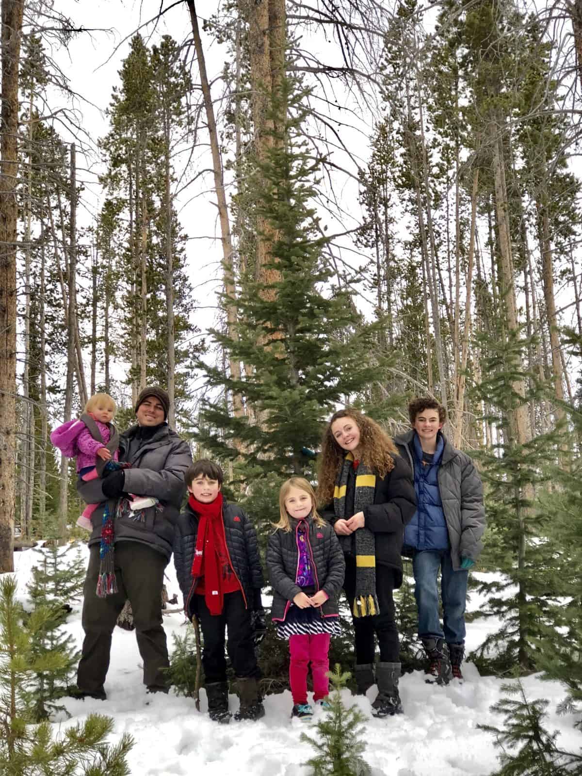 A family stands together amidst snow-covered ground and tall pine trees.
