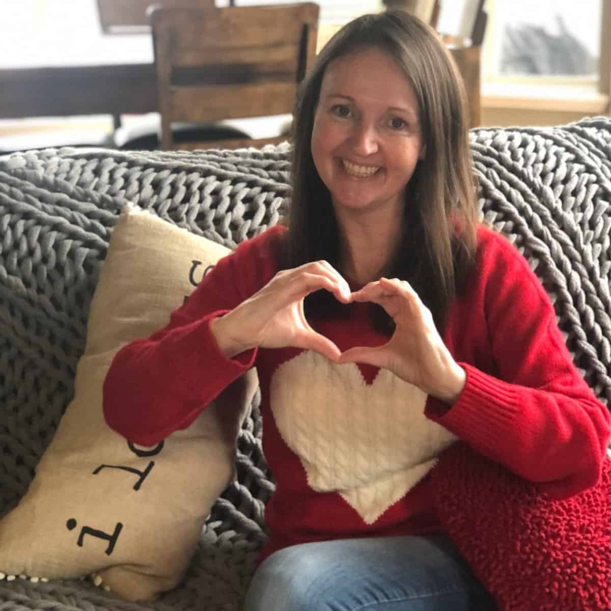 Woman in red sweater making a heart shape with her hands while sitting on a couch.