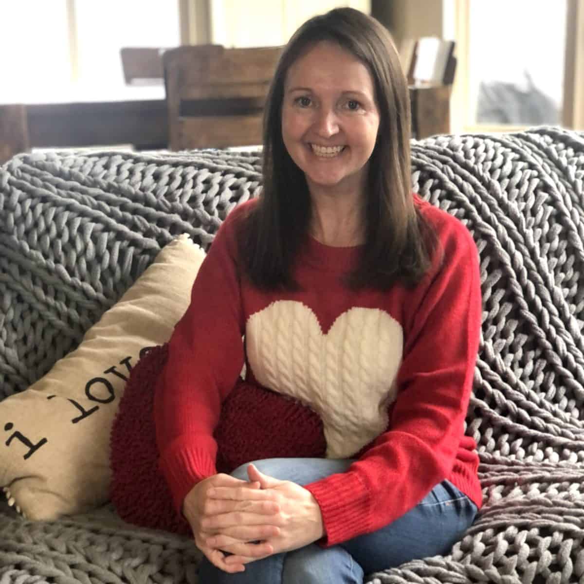 A smiling woman wearing a red sweater with a heart design, seated on a couch with a knitted blanket and a cushion that reads "i love us".