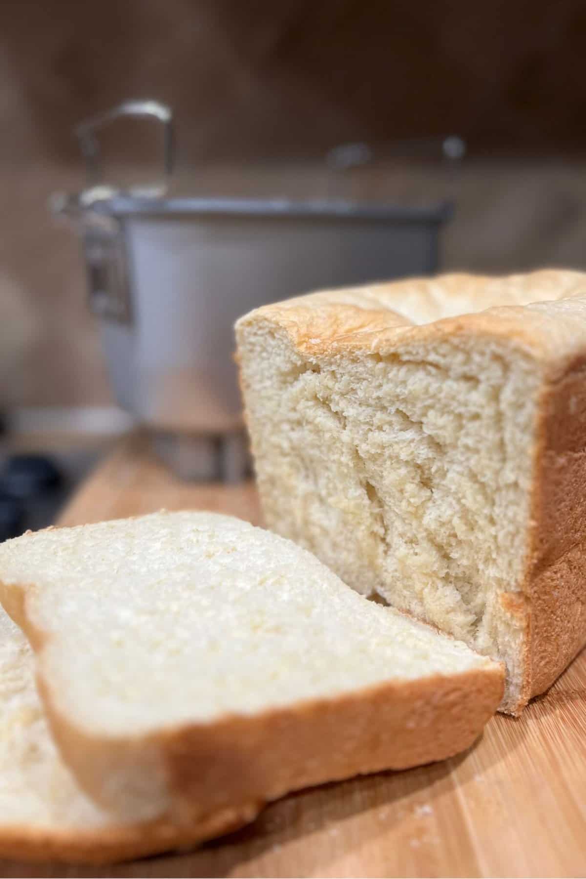 A loaf of white bread made with the Zojirushi Home Bakery Virtuoso Breadmaker, with one slice cut off and placed in the foreground, with a pot on a stove in the background