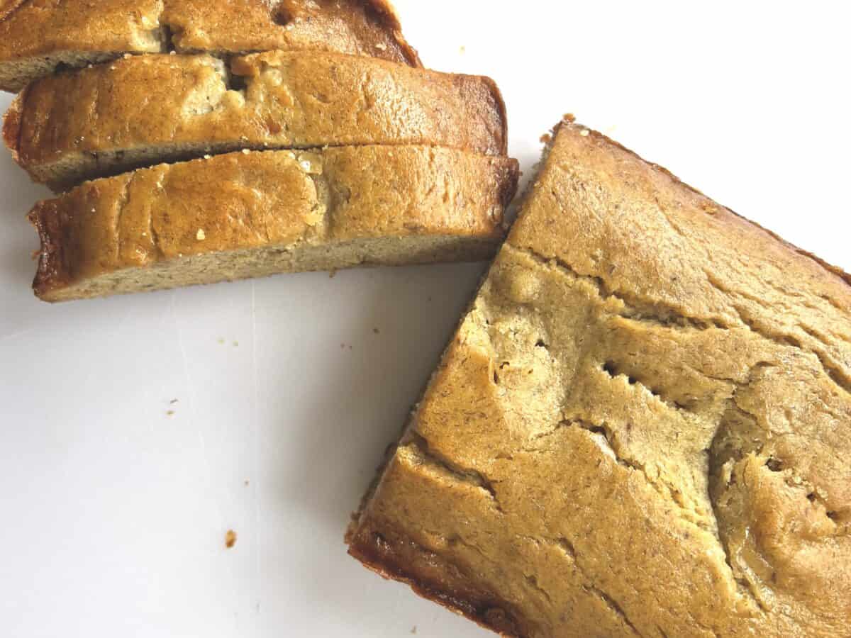 A close-up view of a sliced loaf of freshly baked banana bread resting on a wooden cutting board. The golden brown crust and moist interior of the bread are visible, with small bits of mashed banana.