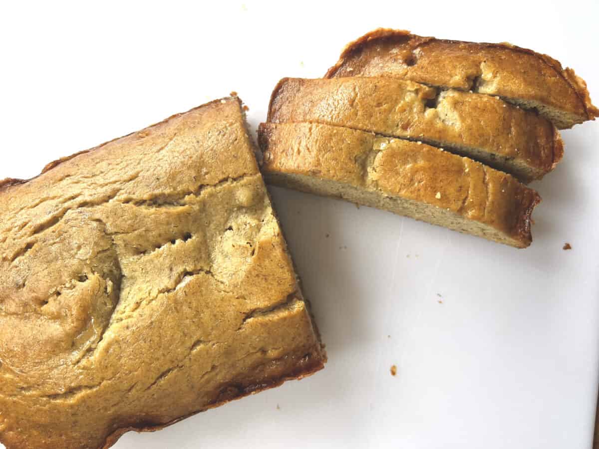 A close-up view of a sliced loaf of freshly baked banana bread resting on a wooden cutting board. The golden brown crust and moist interior of the bread are visible, with small bits of mashed banana