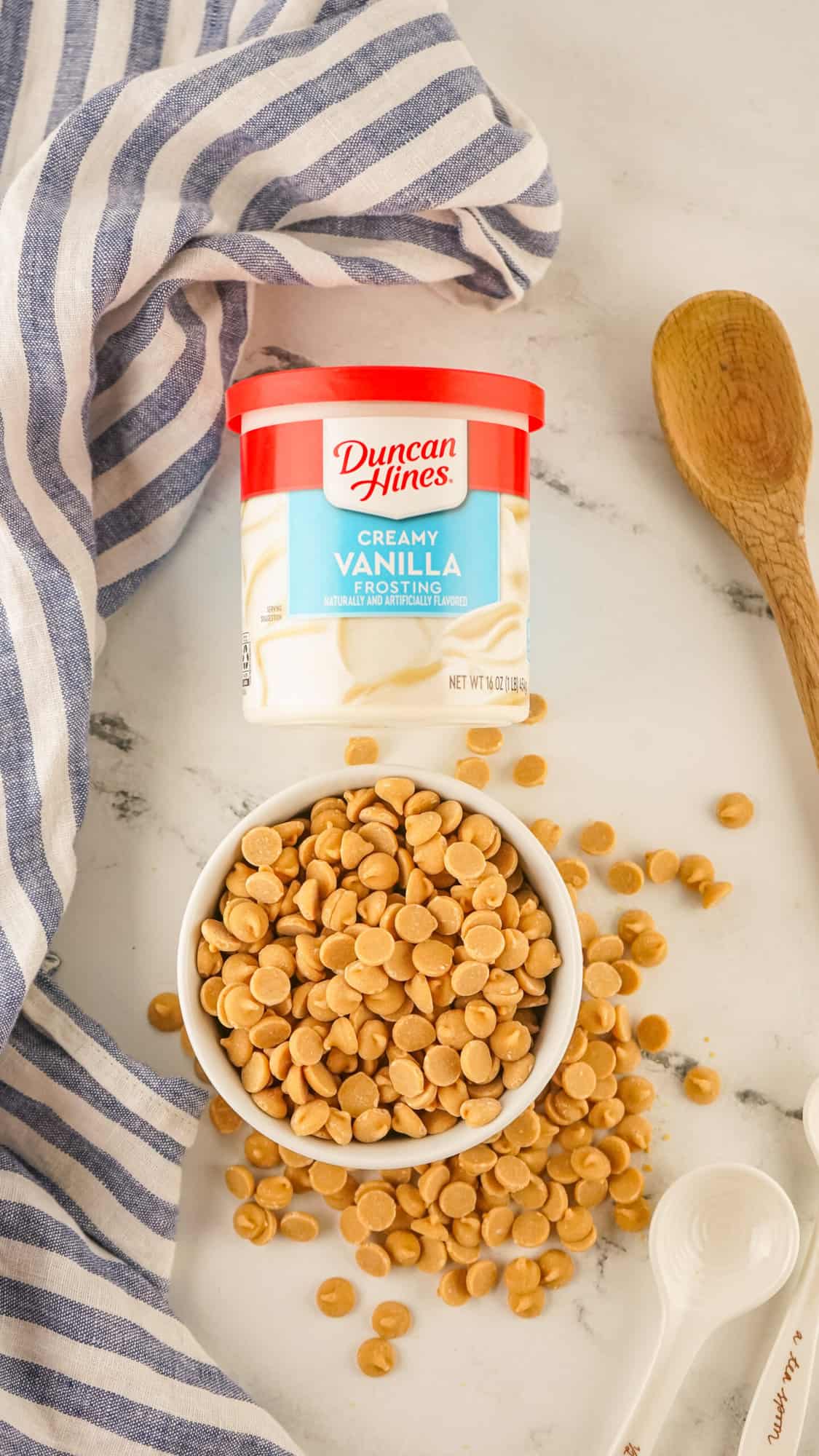 A bowl of butterscotch chips next to a container of duncan hines creamy vanilla frosting and a wooden spoon, with a striped towel in the background.