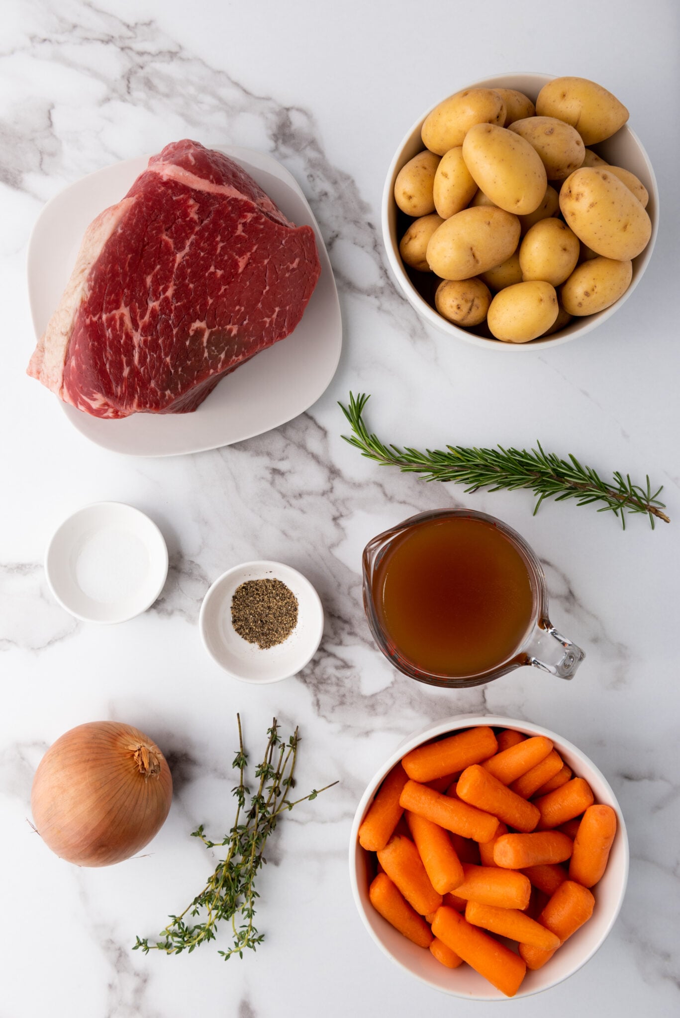 Assorted fresh ingredients laid out, including a rump roast, yellow baby potatoes, baby carrots, an onion, and fresh herbs.