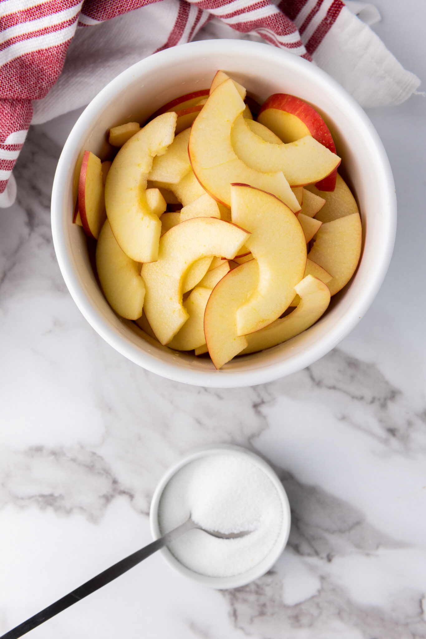 Overhead shot of apple slices in a bowl, sugar being sprinkled, with a spoon