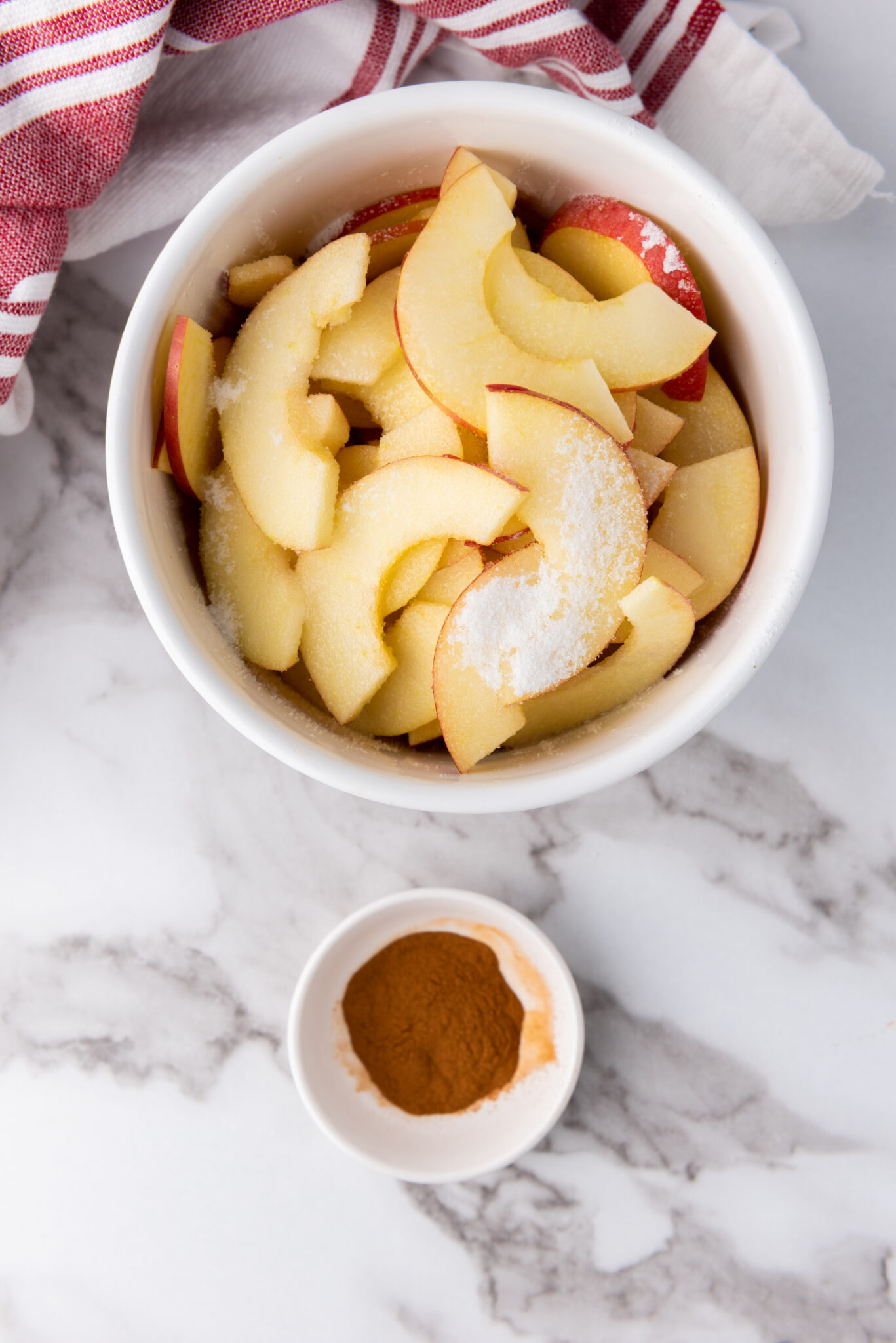 Overhead shot of apple slices in a bowl, sugar and cinnamon being sprinkled