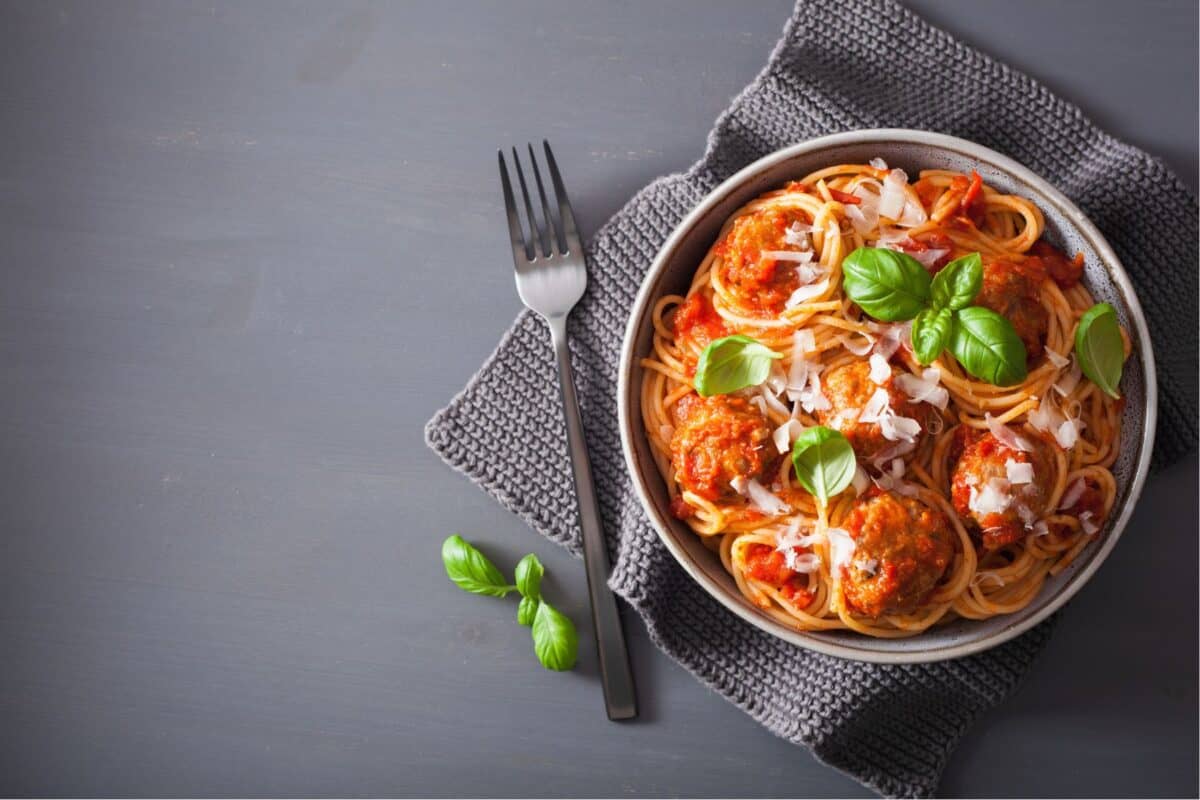 A bowl of spaghetti with meatballs, tomato sauce, grated cheese, fresh basil leaves, and the perfect spaghetti seasoning recipe, accompanied by a fork on a gray napkin.