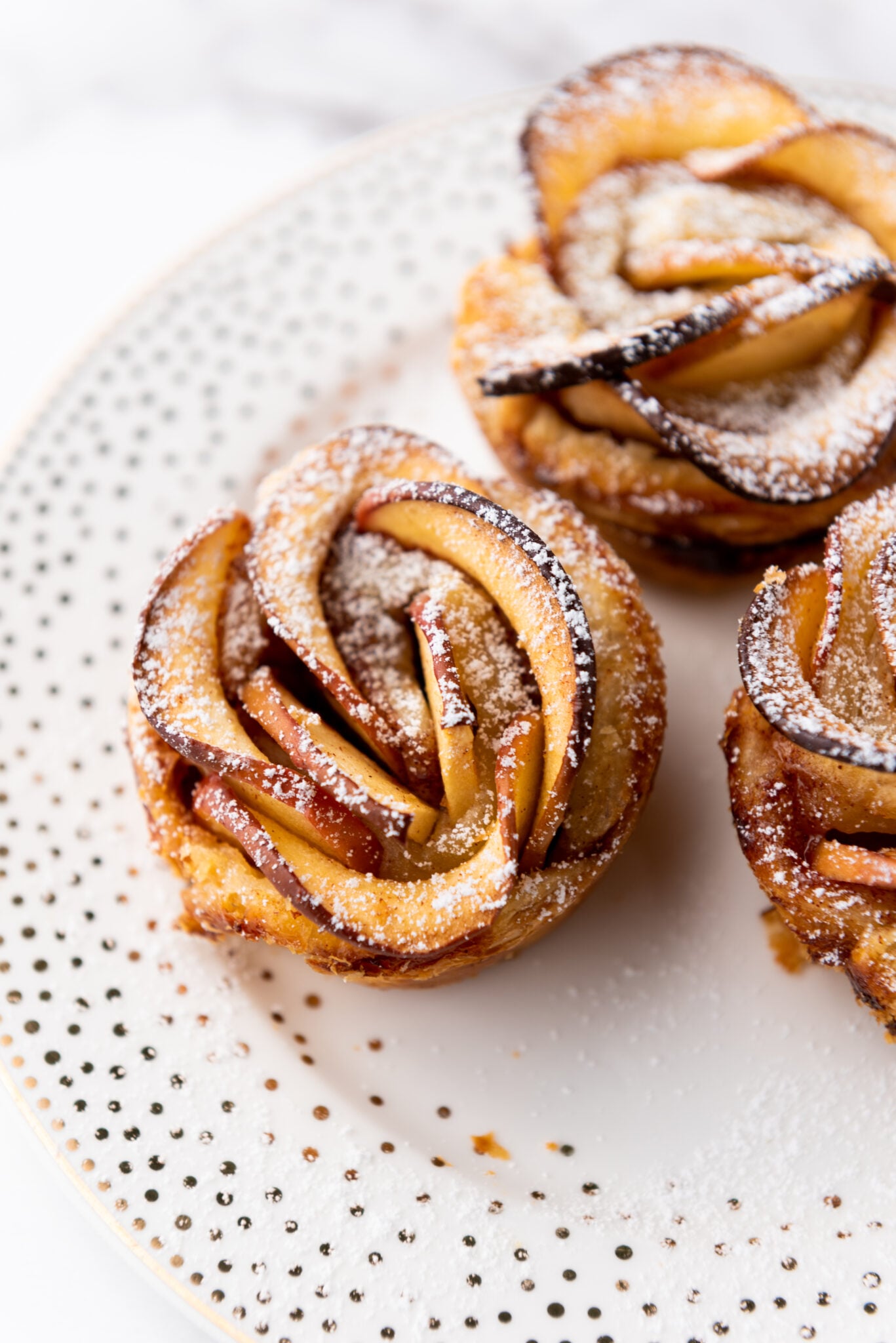 Golden-brown baked apple roses, delicately arranged on a pristine white plate, dusted with powdered sugar, capturing elegance and culinary artistry.
