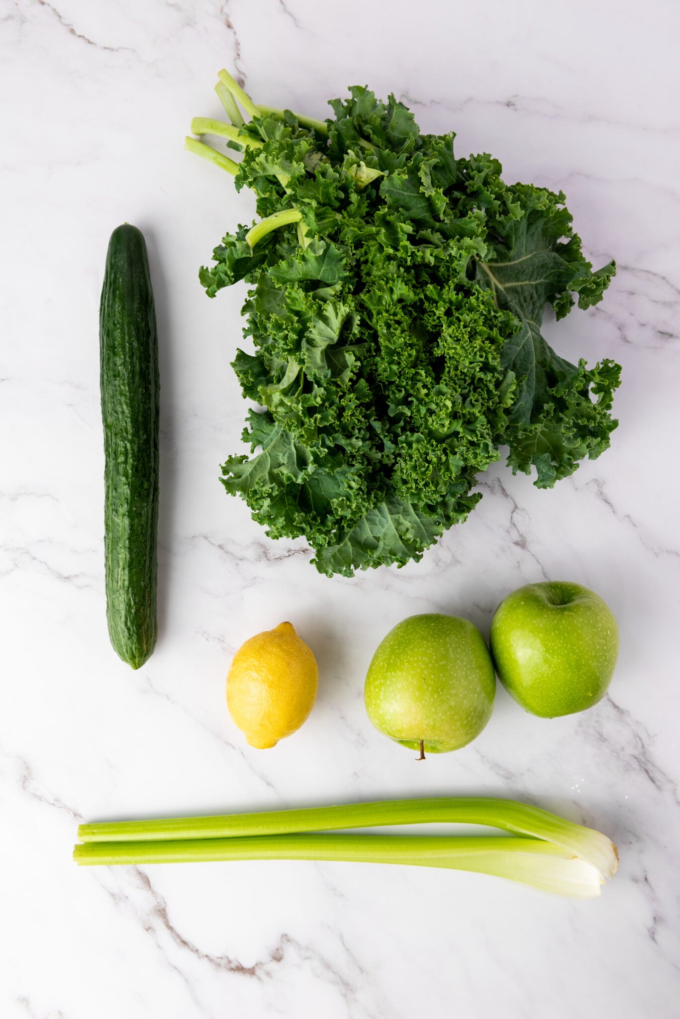 Overhead shot of fresh kale, Granny Smith apples, celery stalks, English cucumber, and a lemon neatly arranged on a white countertop. Hoping for green juice recipes for weight loss.