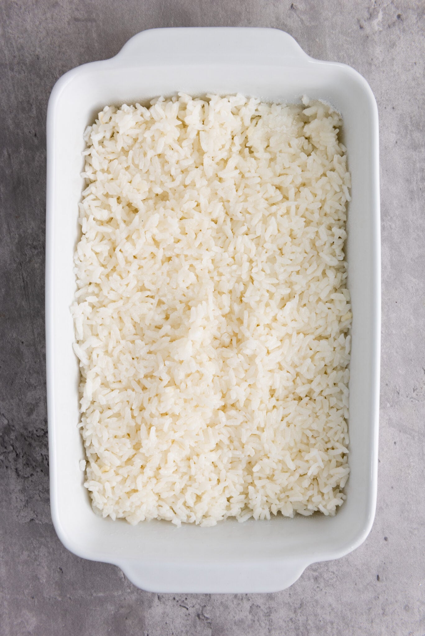 Top view of sushi rice being spread in a glass casserole dish, with sprinklings of vinegar and sugar.