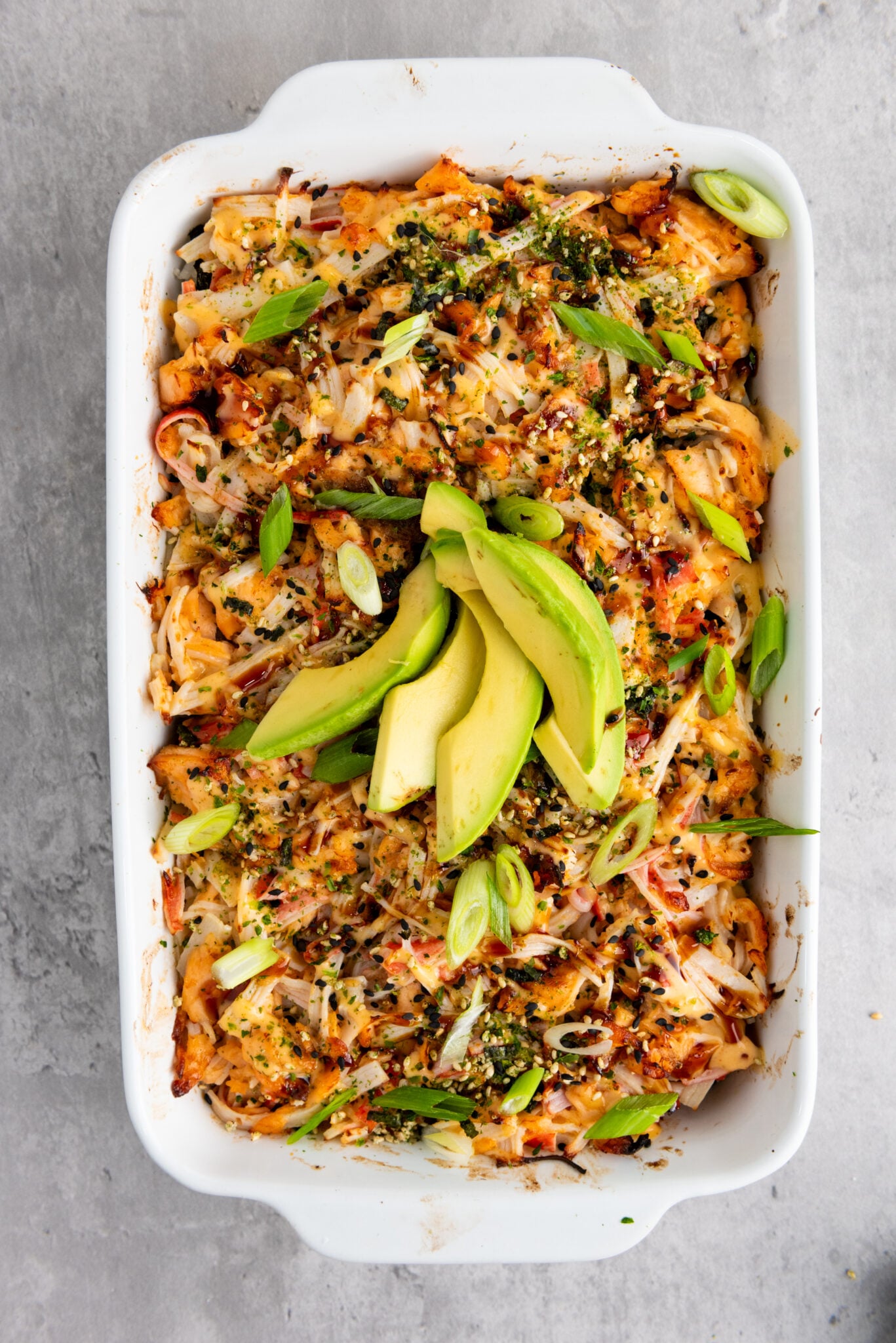 Close-up of sushi bake topped with sauces, seasonings, scallions, and avocado slices.