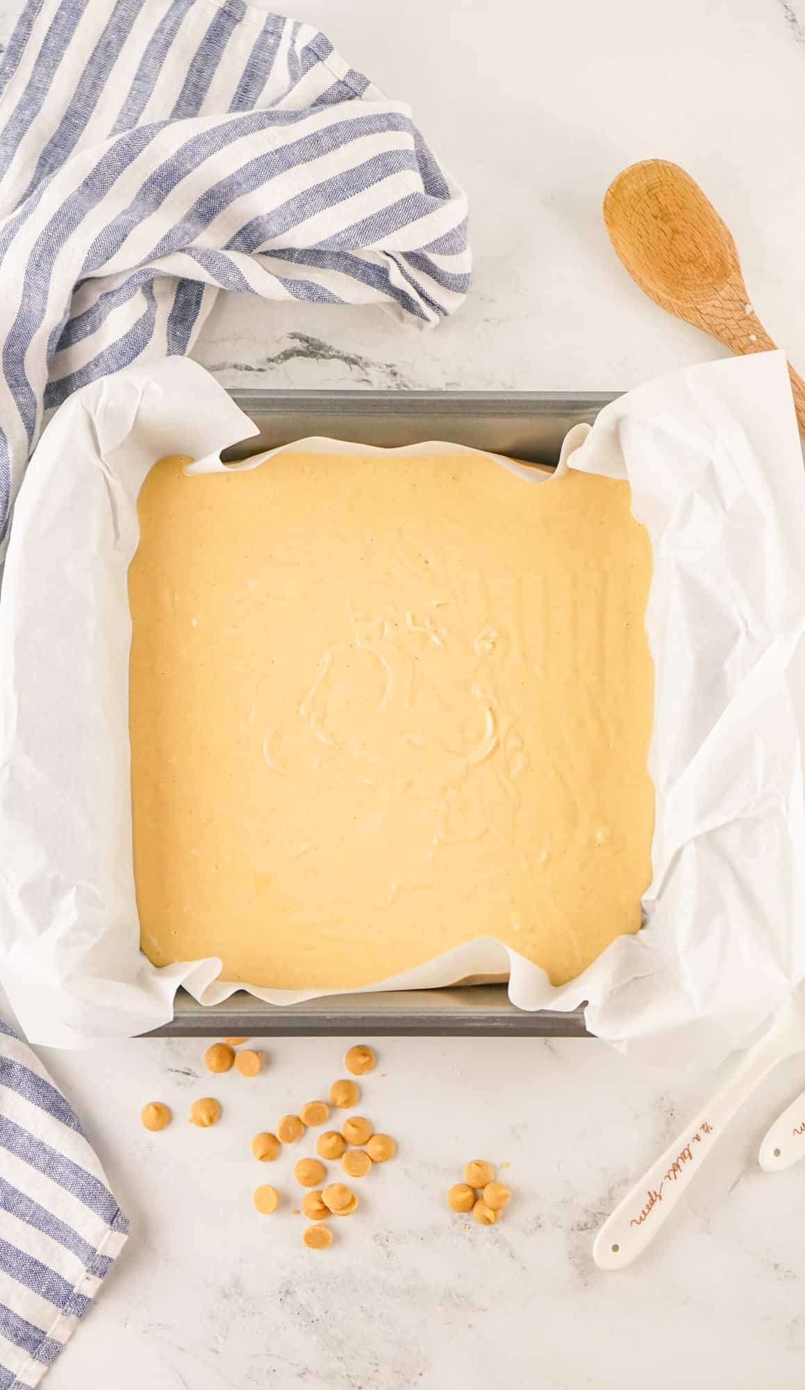 Unbaked blondie batter in a lined baking pan with scattered butterscotch chips and a wooden spoon on a kitchen counter.