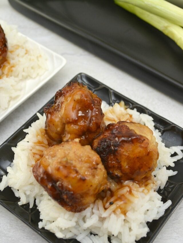 Three teriyaki-glazed meatballs served on a bed of white rice, placed on a black square plate. A plate with green vegetables is partially visible in the background.