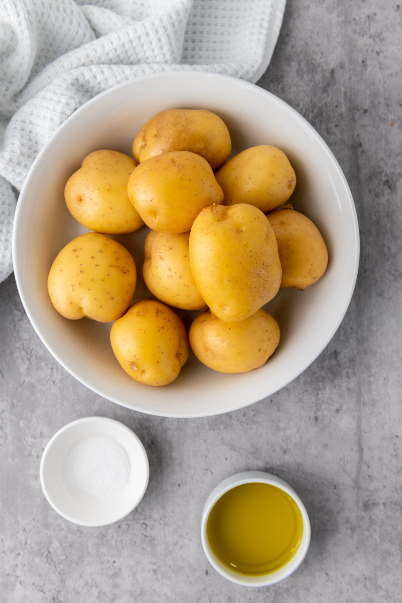 A bowl of raw yellow potatoes on a light gray surface with a small bowl of salt and a cup of oil nearby, ready for an Accordion Potatoes recipe using the Air Fryer.