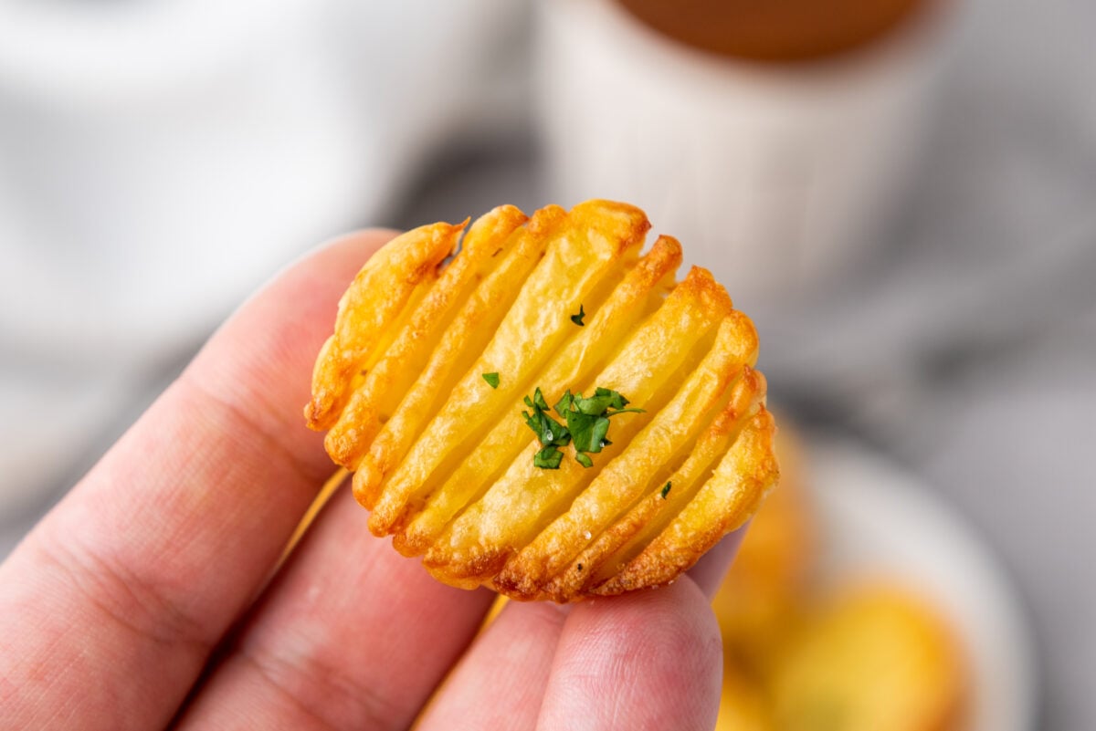 A close-up of a hand holding a small, golden-brown accordion potato stack garnished with parsley.