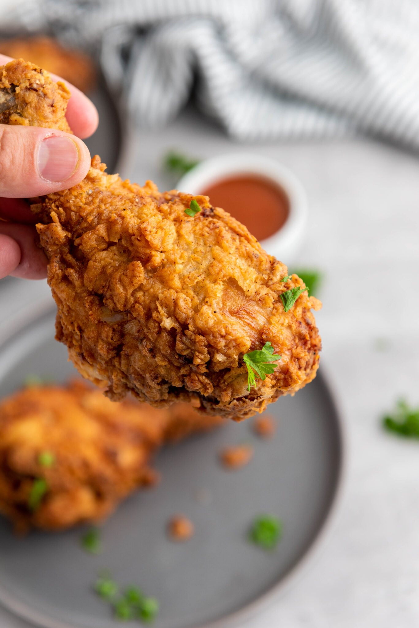 A person holding a crispy chicken thigh with more pieces on a plate in the background.