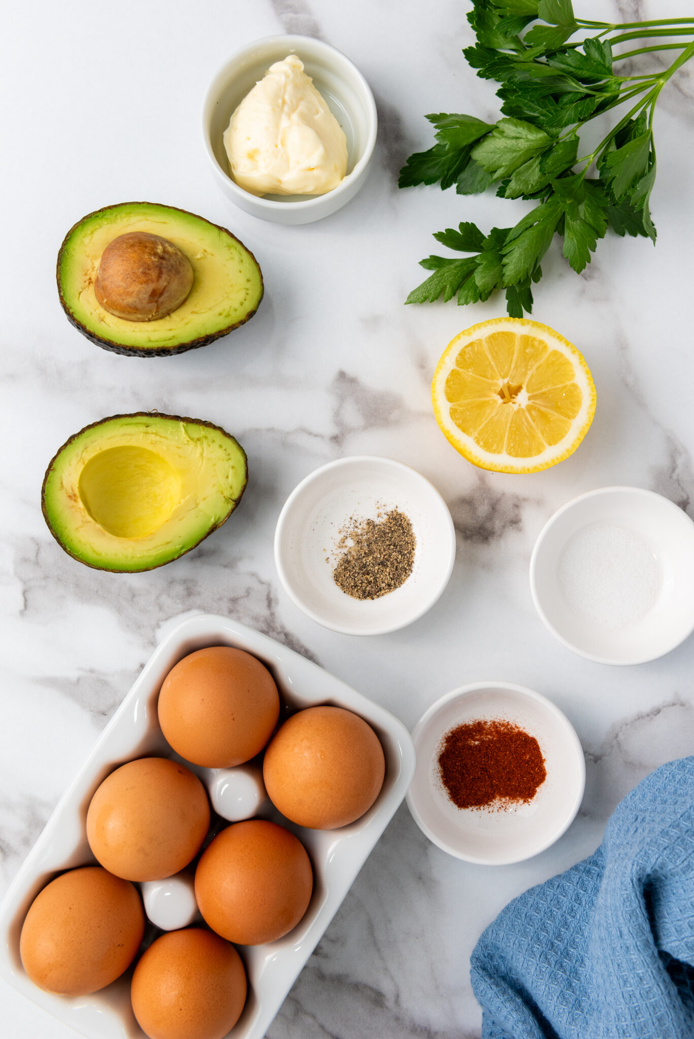 A variety of ingredients for making avocado deviled eggs are displayed on a light-colored wooden table. In the center is a halved avocado with a visible pit. Next to the avocado are six whole eggs. Other ingredients scattered around the table include a small bowl of sour cream, a halved lemon, a white container with a black cap, a small black pepper mill, and a small green container with a red lid. Fresh parsley sprigs and a red paprika powder shaker are positioned near the avocado half.