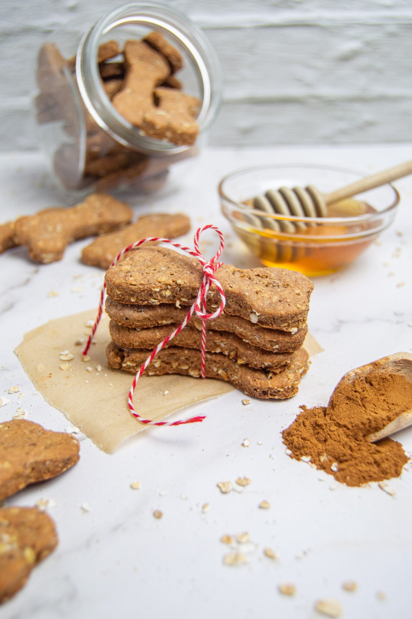 A stack of homemade whole wheat dog treats with a red and white string, next to a jar of honey and scattering of oats and cinnamon.